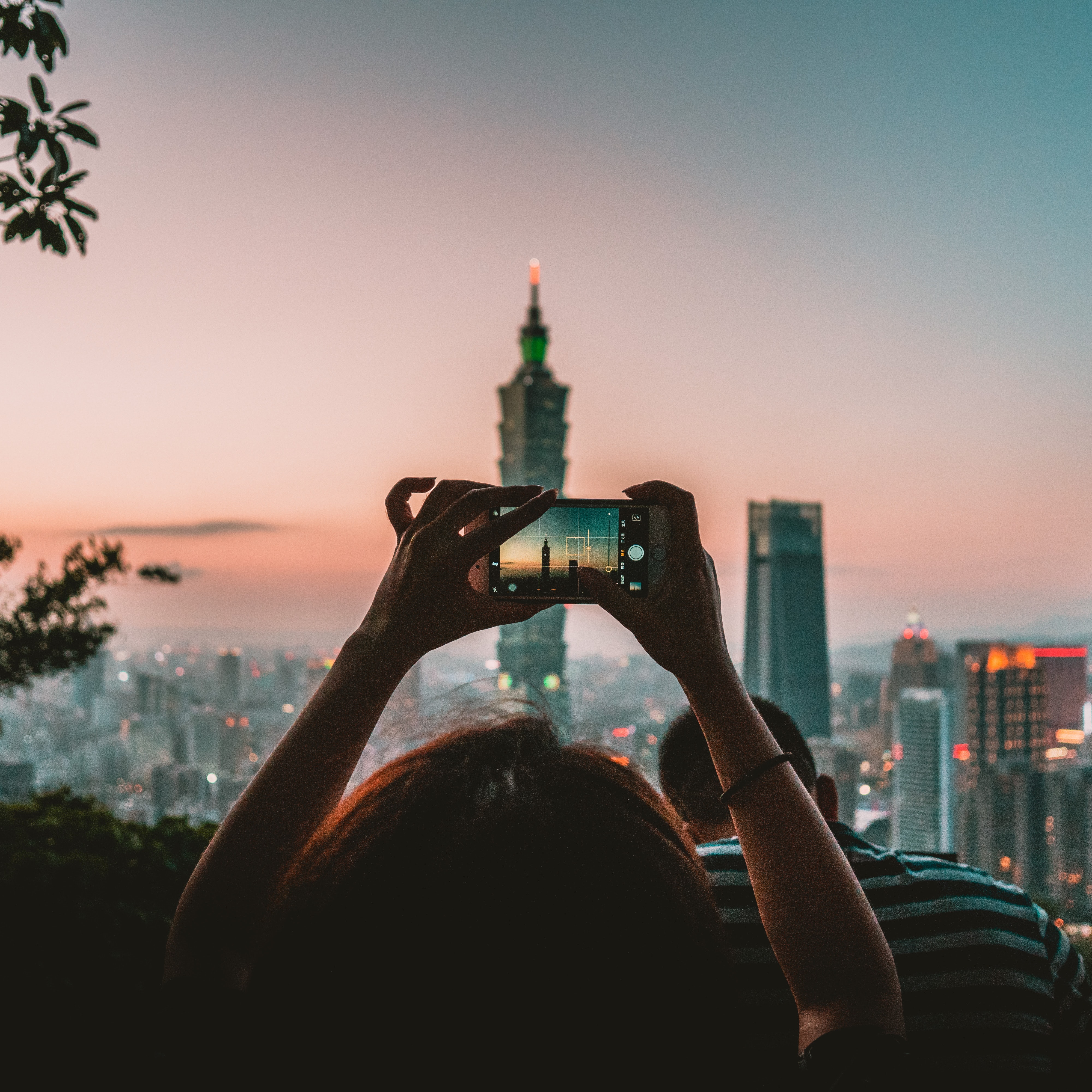 A young ladie holding a smartphone to take a picture of a big building with a sunset on the background